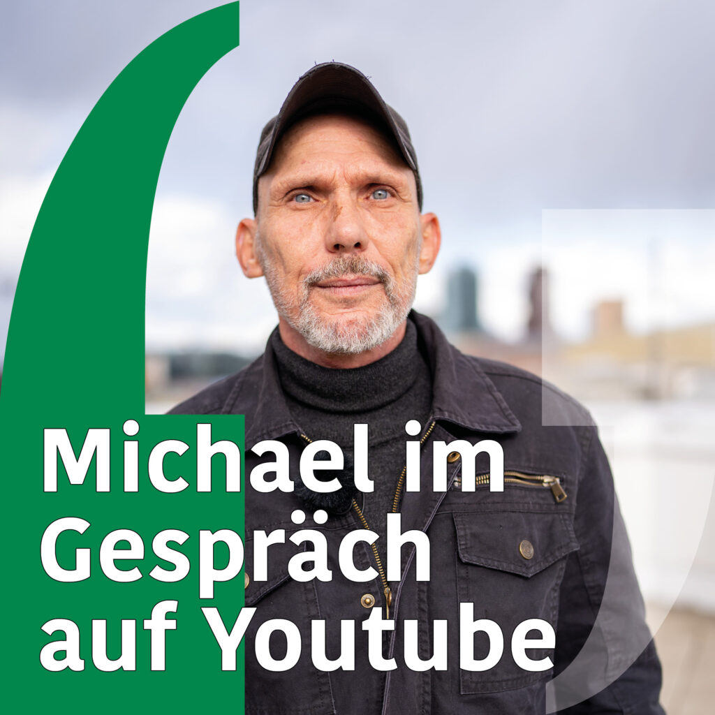 Foto eines ca. 50-60 Jährigen weißen Mannes mit blauen Augen und Kinnbart. Er trägt eine schwarze Kappe und schwarze Jacke. Text: "Michael im Gespräch auf Youtube".