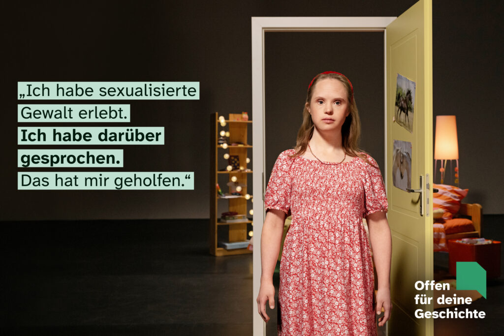 Motiv der Kampagne "Offen für deine Geschichte": Eine junge Frau mit Down-Syndrom steht im Zentrum des Fotos. Sie ist blond und hat ein rot-weiß geblümtes Kleid an. Im Hintergrund eine Theaterkulisse mit geöffneter Gelber Tür. Dahinter ist die Einrichtung eines Kinderzimmers zu erkennen. Text links: "Ich habe sexualisierte Gewalt erlitten. Ich habe darüber gesprochen. Das hat mir geholfen." Rechts unten Logo der Kampagne