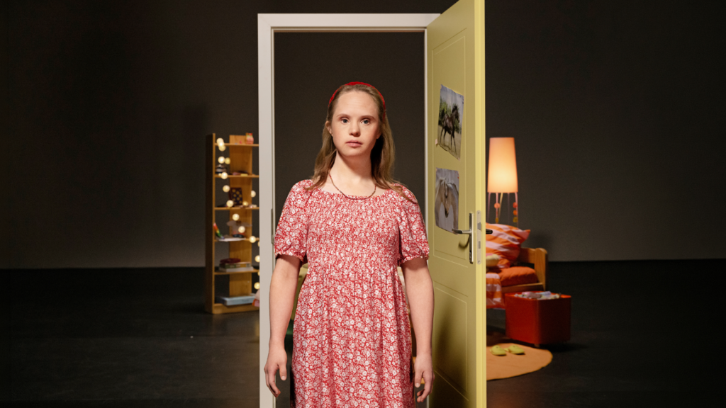 Motiv der Kampagne "Offen für deine Geschichte": Eine junge Frau mit Down-Syndrom steht im Zentrum des Fotos. Sie ist blond und hat ein rot-weiß geblümtes Kleid an. Im Hintergrund eine Theaterkulisse mit geöffneter Gelber Tür. Dahinter ist die Einrichtung eines Kinderzimmers zu erkennen.