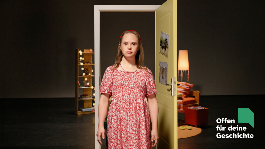 Motiv der Kampagne "Offen für deine Geschichte": Eine junge Frau mit Down-Syndrom steht im Zentrum des Fotos. Sie ist blond und hat ein rot-weiß geblümtes Kleid an. Im Hintergrund eine Theaterkulisse mit geöffneter Gelber Tür. Dahinter ist die Einrichtung eines Kinderzimmers zu erkennen. Rechts unten Logo der Kampagne.