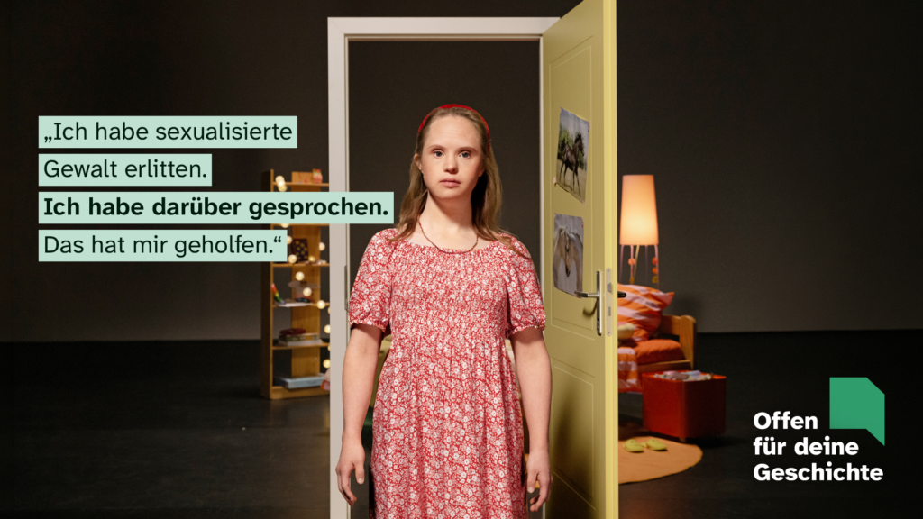 Motiv der Kampagne "Offen für deine Geschichte": Eine junge Frau mit Down-Syndrom steht im Zentrum des Fotos. Sie ist blond und hat ein rot-weiß geblümtes Kleid an. Im Hintergrund eine Theaterkulisse mit geöffneter Gelber Tür. Dahinter ist die Einrichtung eines Kinderzimmers zu erkennen. Text links: "Ich habe sexualisierte Gewalt erlitten. Ich habe darüber gesprochen. Das hat mir geholfen." Rechts unten Logo der Kampagne