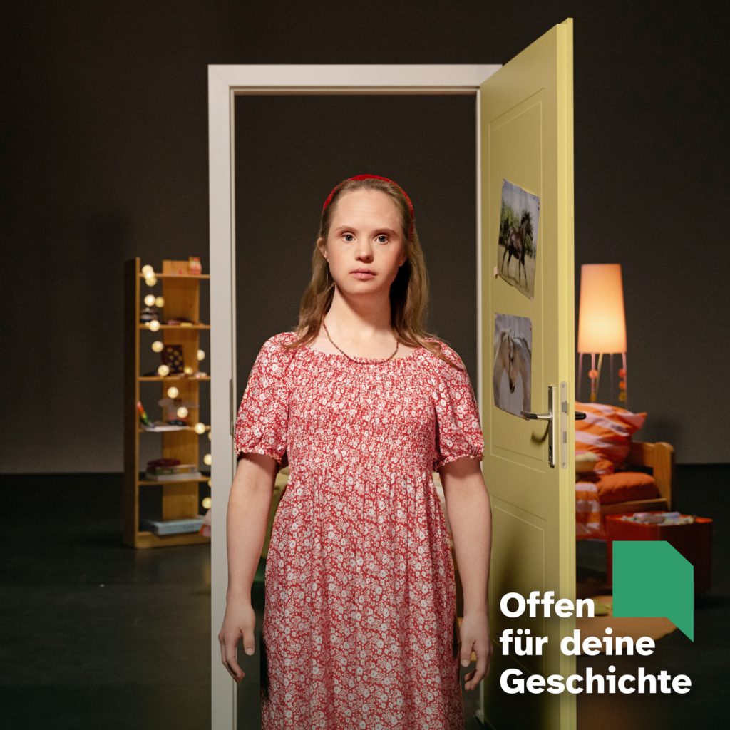 Motiv der Kampagne "Offen für deine Geschichte": Eine junge Frau mit Down-Syndrom steht im Zentrum des Fotos. Sie ist blond und hat ein rot-weiß geblümtes Kleid an. Im Hintergrund eine Theaterkulisse mit geöffneter Gelber Tür. Dahinter ist die Einrichtung eines Kinderzimmers zu erkennen. Unten rechts Logo "Offen für deine Geschichte"